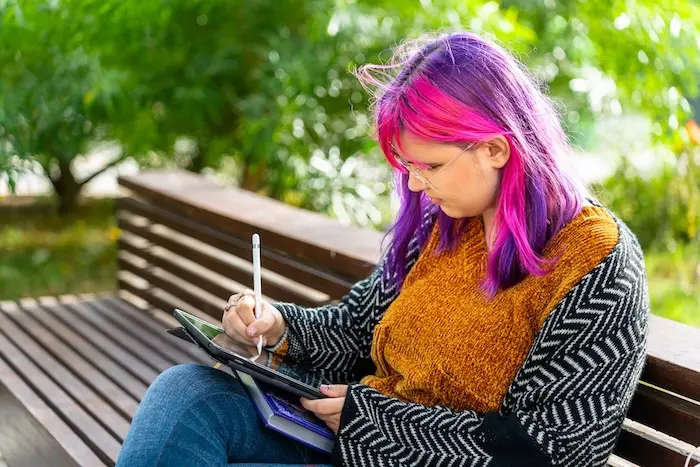 A young woman with pink hair using a stylus to create digital art on a tablet while outdoors, illustrating creative career paths like graphic design and illustration for adults with autism.